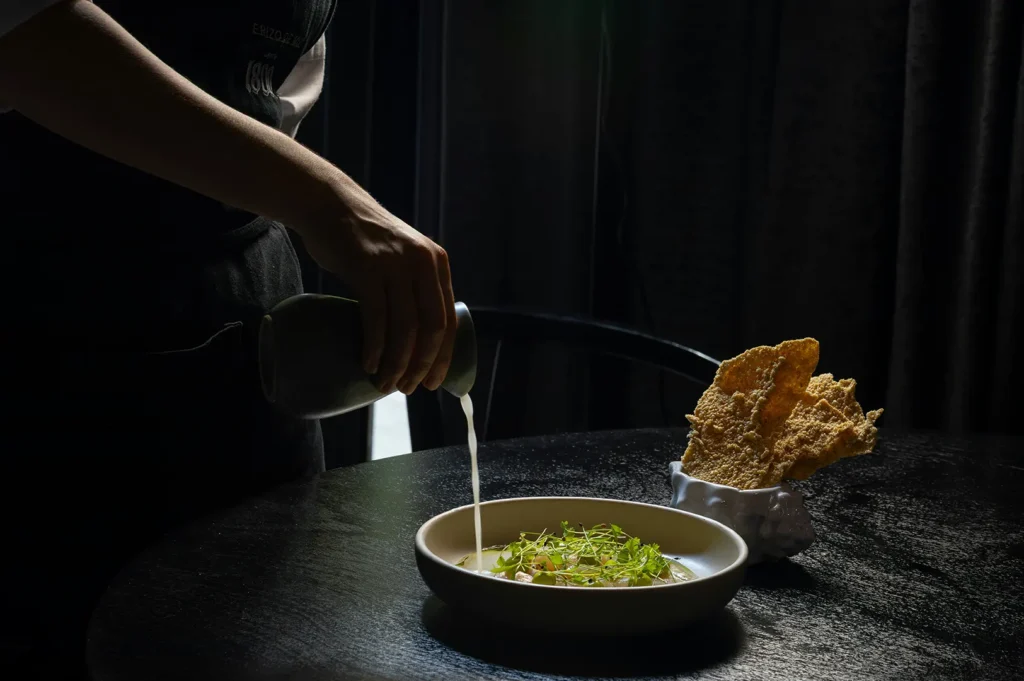 Dark mood commercial food photography of a chef pouring sauce over a plated dish with crisp side crackers on a black table.