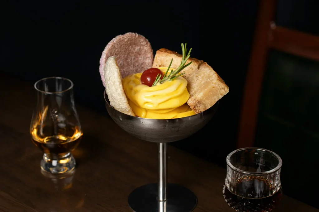 Dark moody food photography of a gourmet dessert in a metal glass with side glasses of drinks.