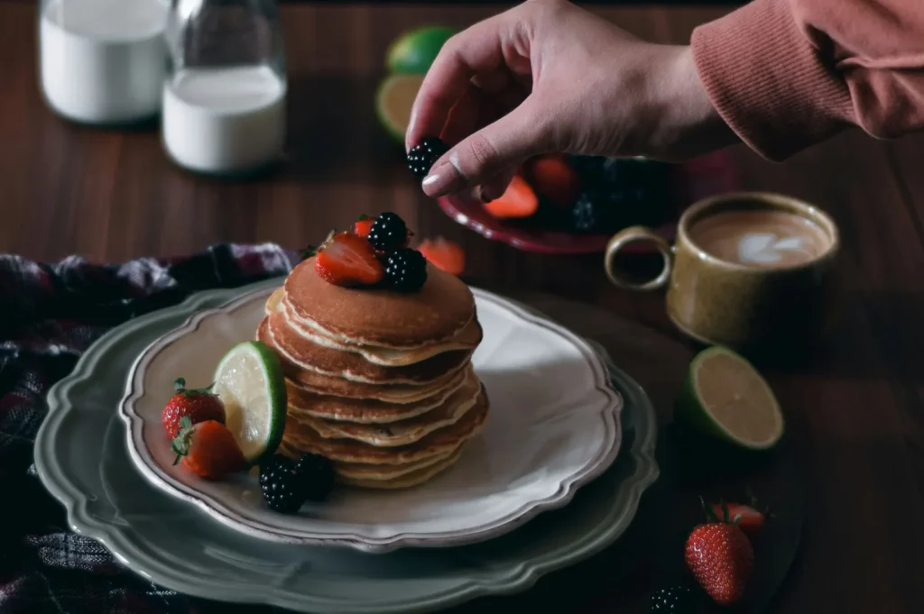 Hand placing a blackberry on top of a stack of pancakes styled with berries and coffee in a dark aesthetic food photo.