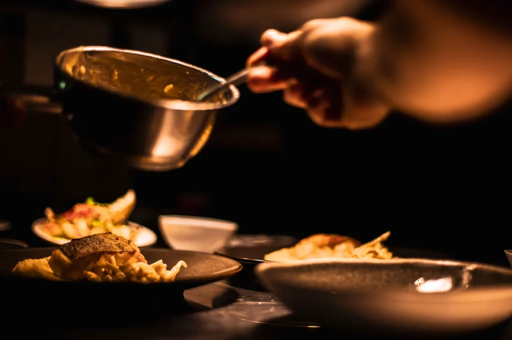 Chef plating gourmet dish in a dark professional restaurant kitchen under moody low-key lighting.