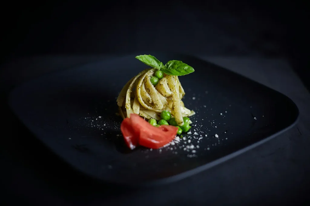 A plate of gourmet pesto pasta with green peas and tomato on a dark moody black background.