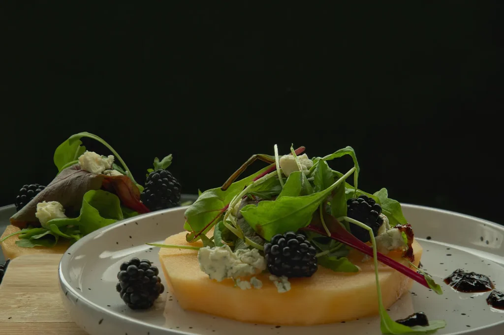 A plated dish of sliced melon with blackberries, blue cheese, and arugula.