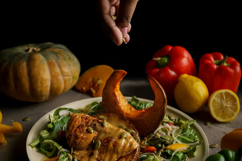 Dark-styled commercial food photography of a healthy salad with roasted pumpkin and chicken, garnished with seeds against a black background.