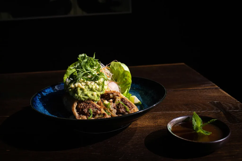 Professional low-key food styling of a signature meat dish with herbs and dipping sauce against a dark background.