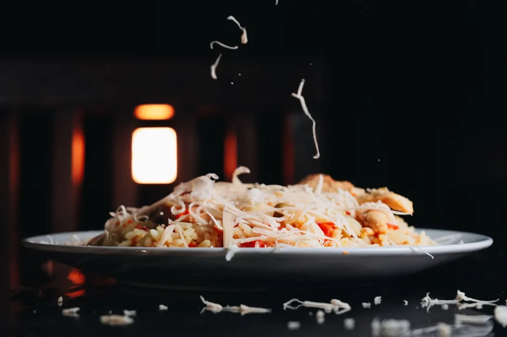 Dark moody food photography of a plate of rice dish being topped with falling shredded cheese against a black background.