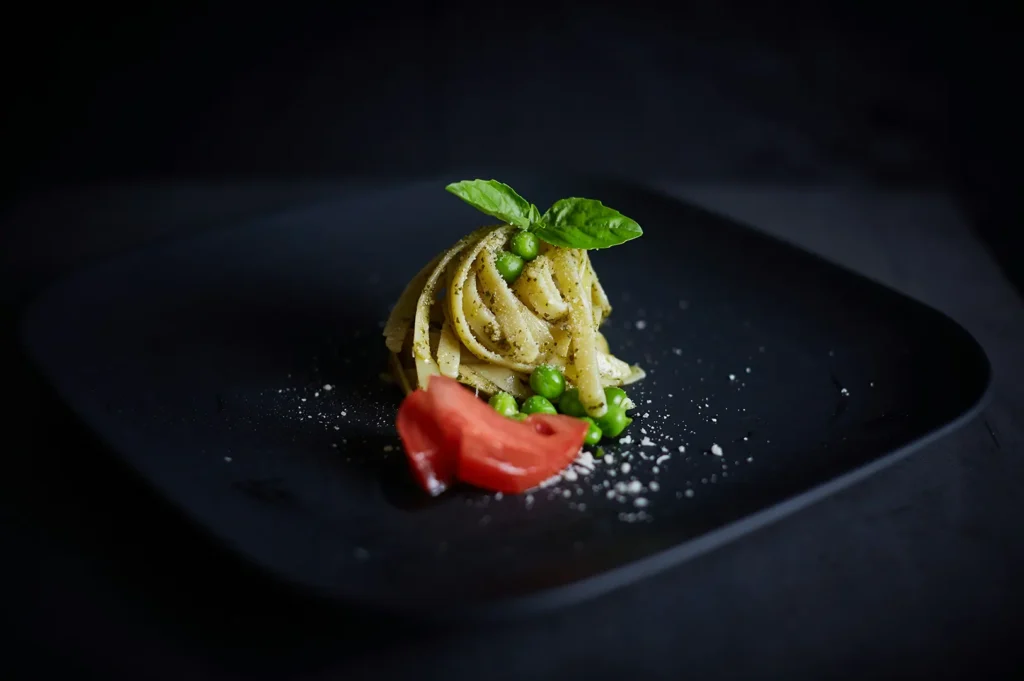 Dark moody food photography of gourmet pasta plated with green peas, tomato garnish, and basil on a black plate.