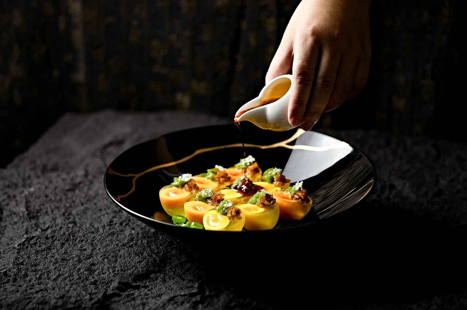 Chef pouring sauce over a plated dish in a dark food photography setting.