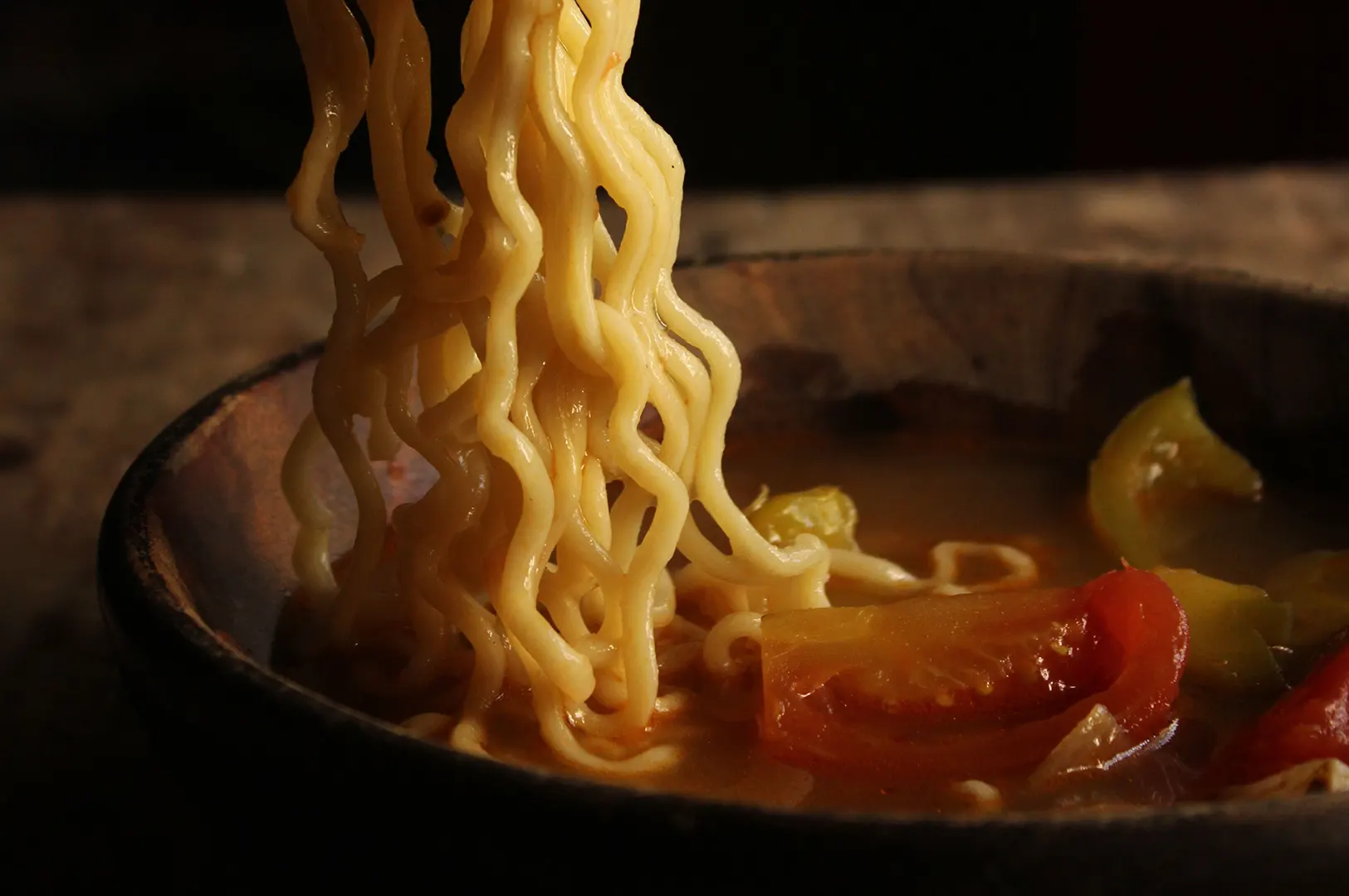 Dark moody food photography of noodles being lifted from a bowl of clear broth with tomatoes.