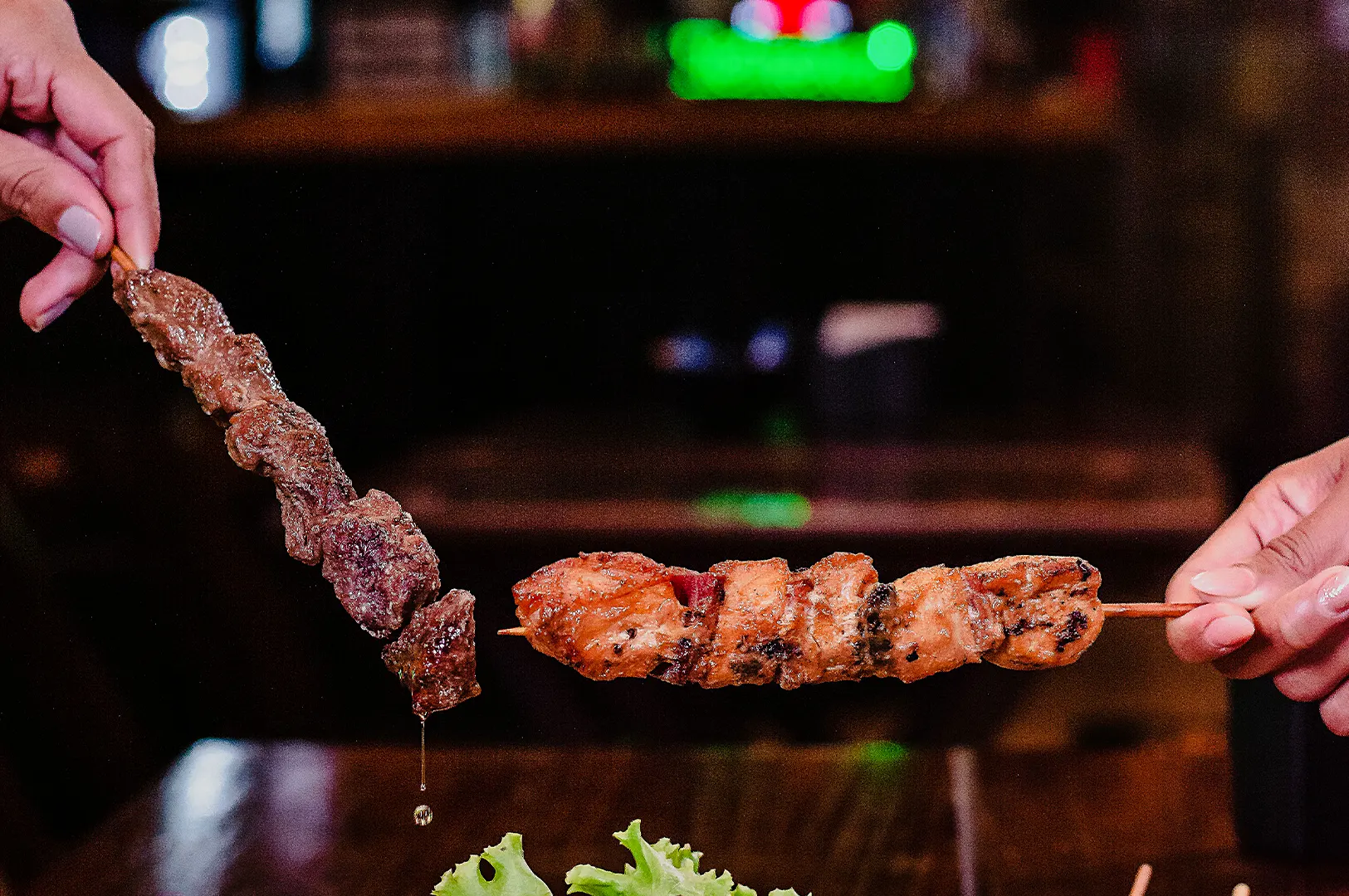 Dark moody food photography of two hands holding sharing skewers against a dark background.