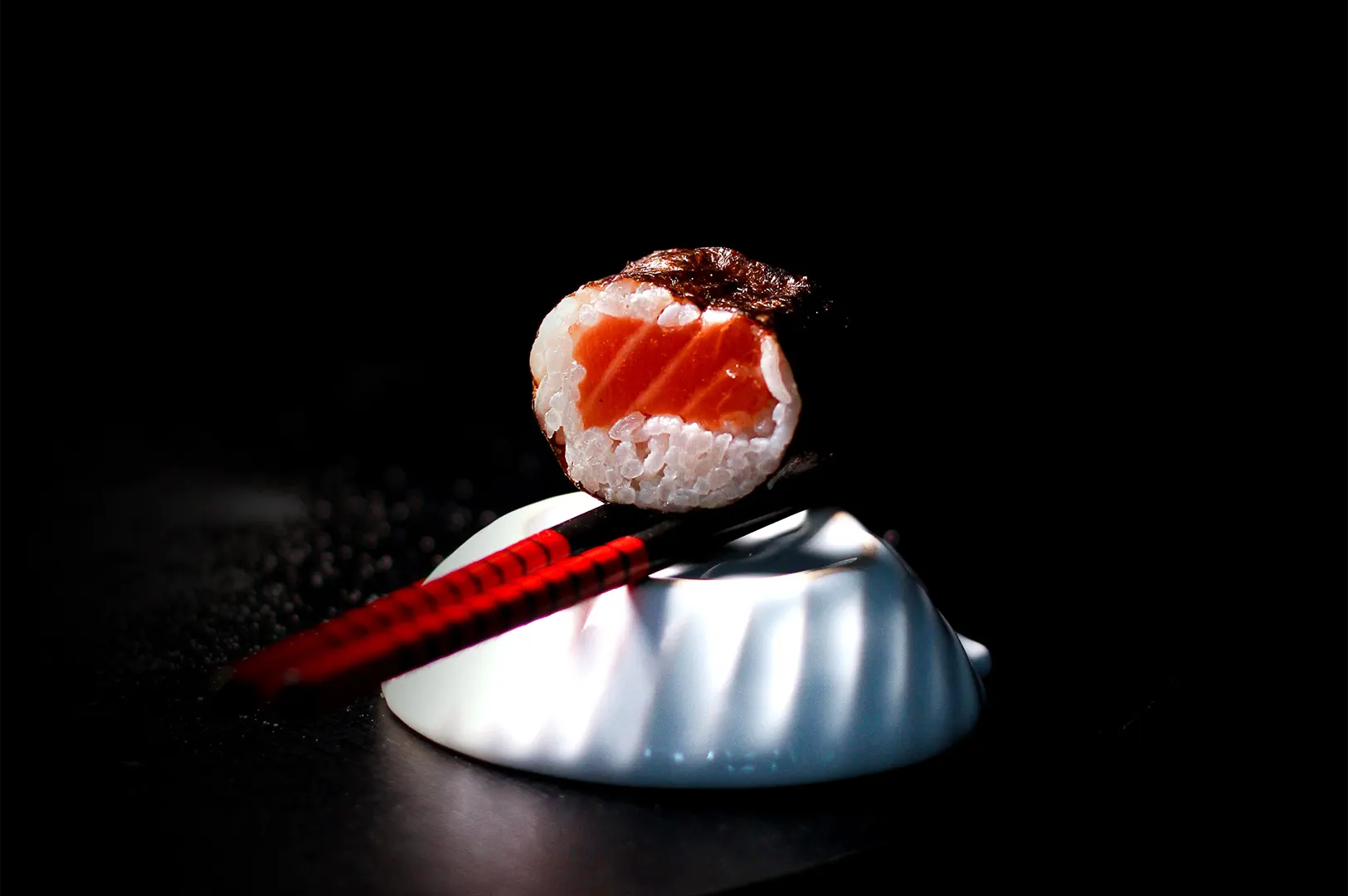 Dark and moody food photography of salmon maki sushi on a white dish with red chopsticks.