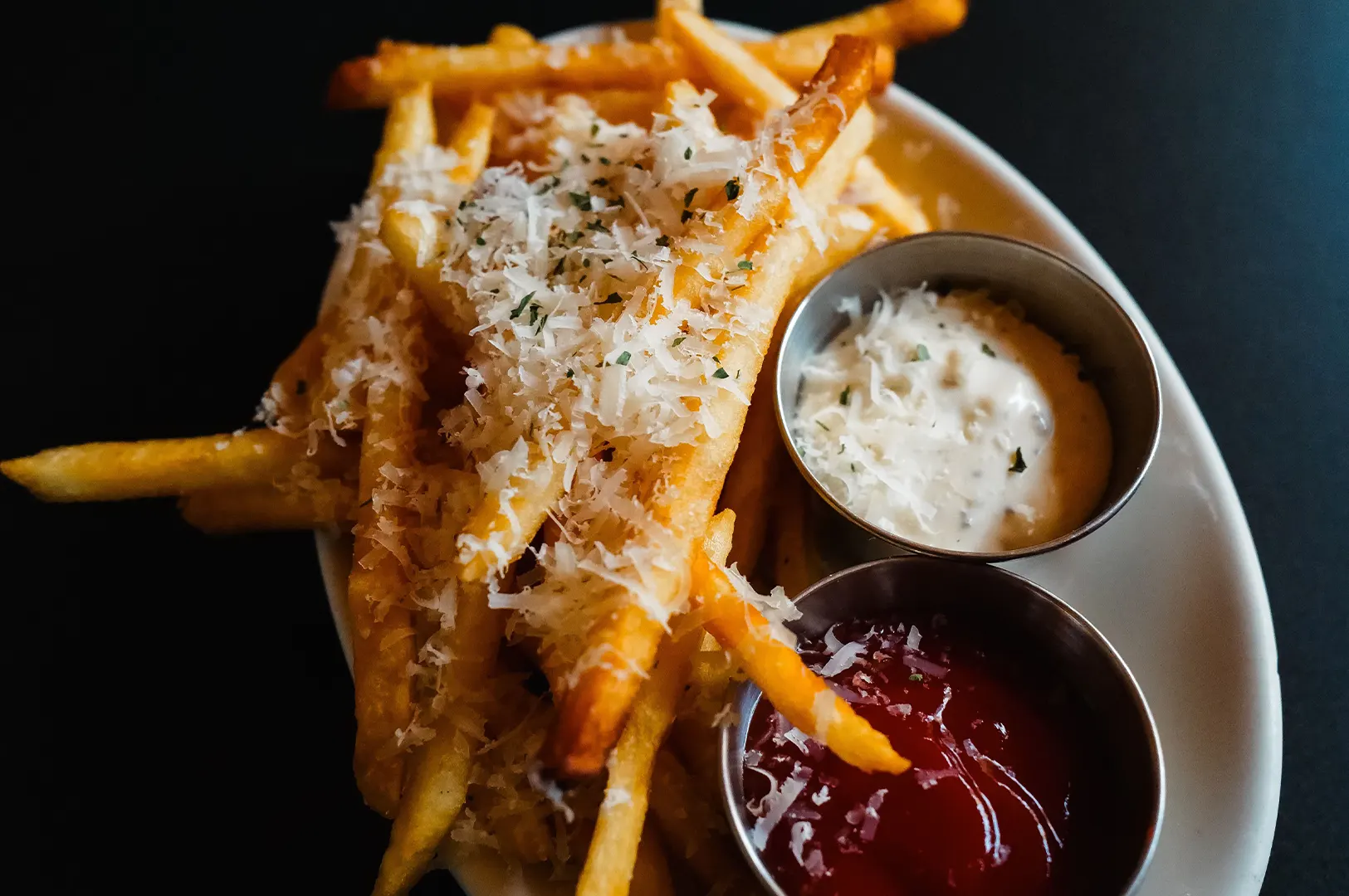 Close-up of crispy truffle fries topped with grated cheese, served with dipping sauces on a dark background.