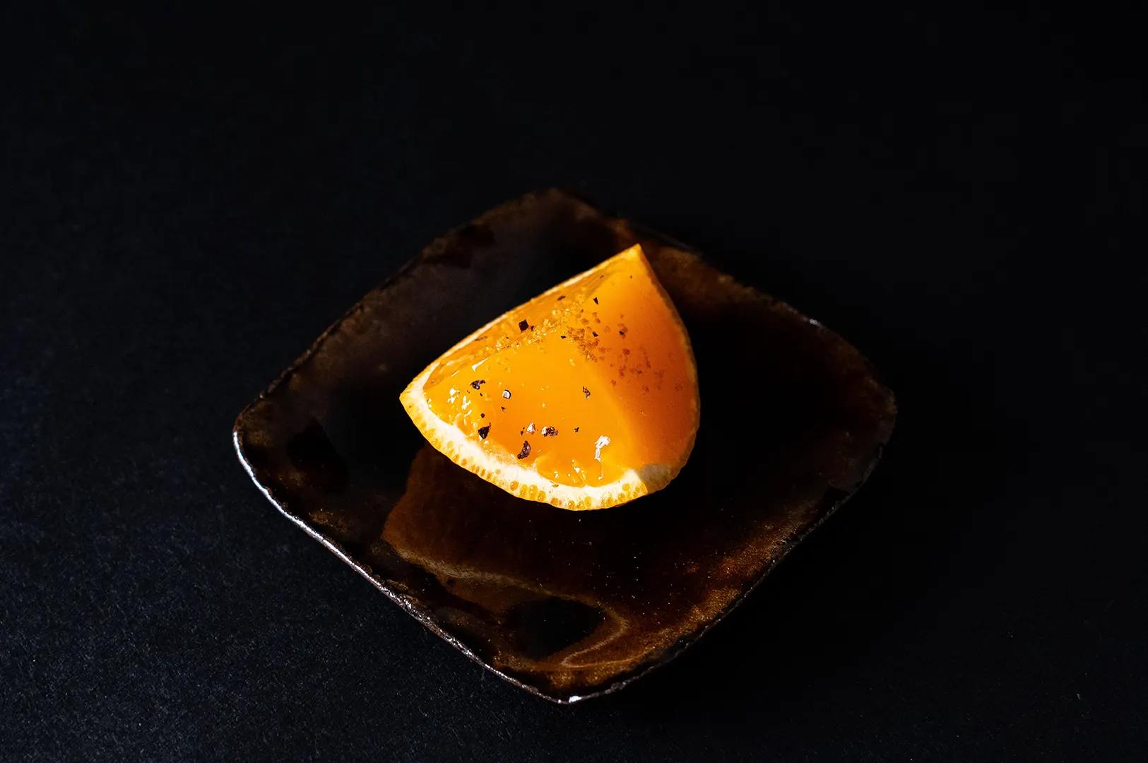 Dark moody food photography of a fresh orange wedge on a dark square plate against a black background.