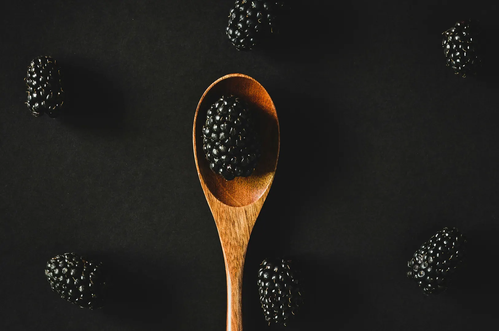 Dark moody food photography of fresh blackberries on a wooden spoon against a black background.