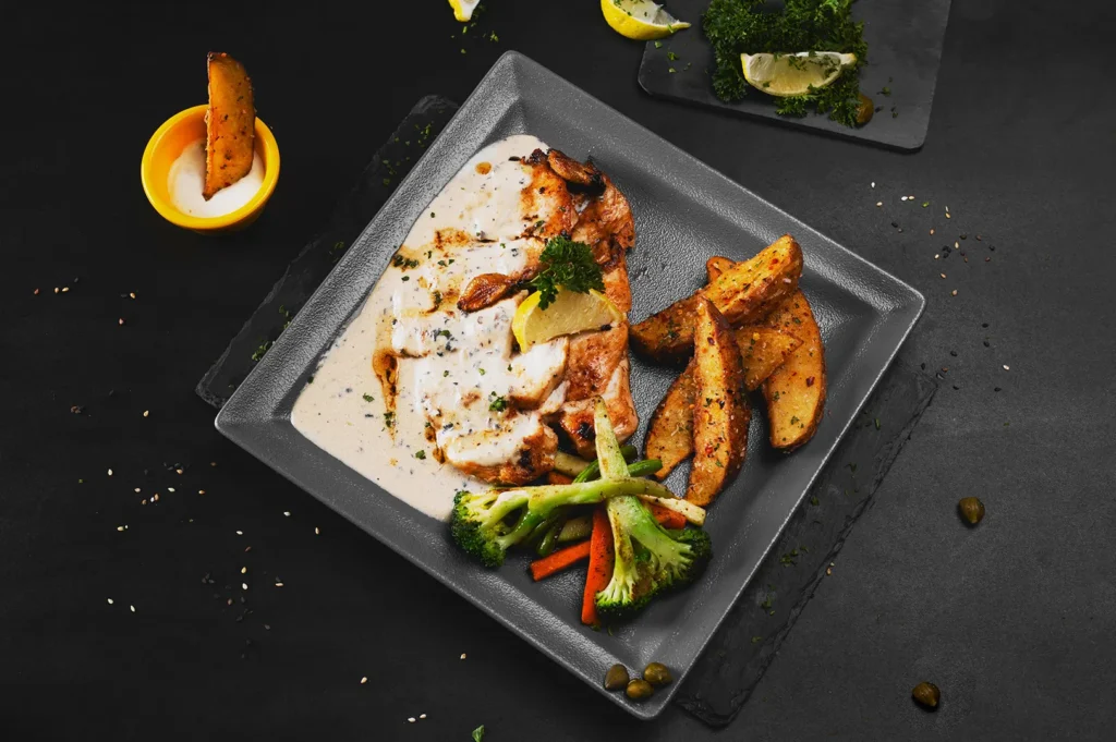 Plated chicken with white sauce, potato wedges, and vegetables on a dark background.