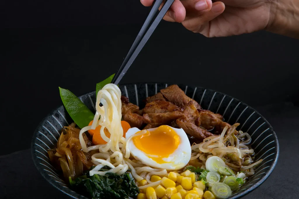 Hand using chopsticks lifting noodles from a bowl of ramen with a soft-boiled egg against a dark background.