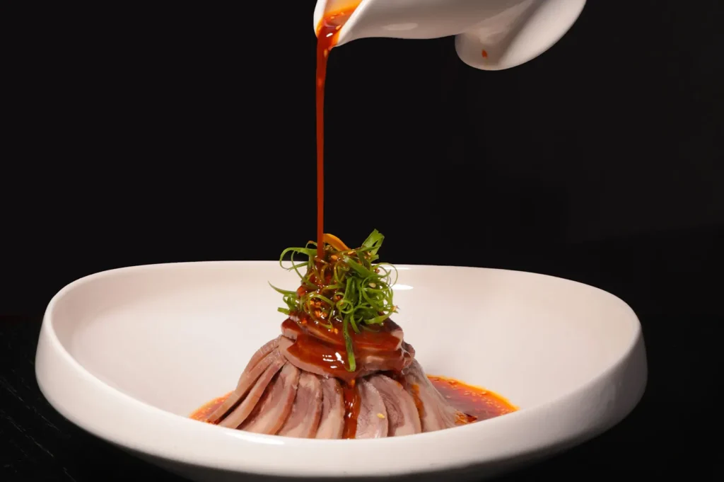 Sauce being poured over a sliced meat dish garnished with scallions in a white bowl.