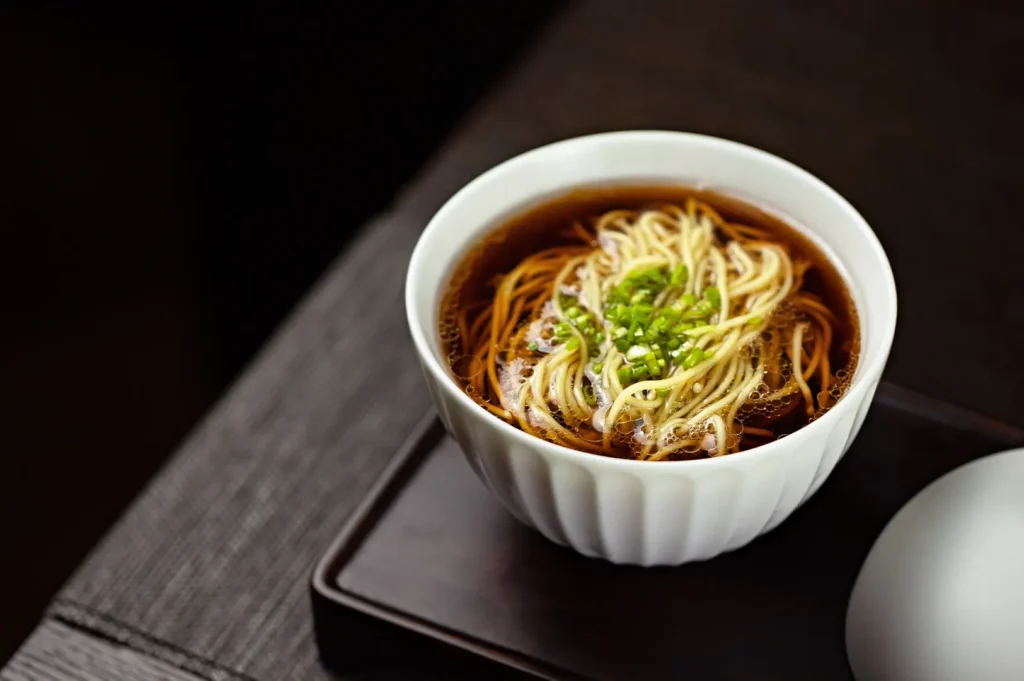 Dark and moody food photography of a bowl of clear soup and noodles with green garnish.