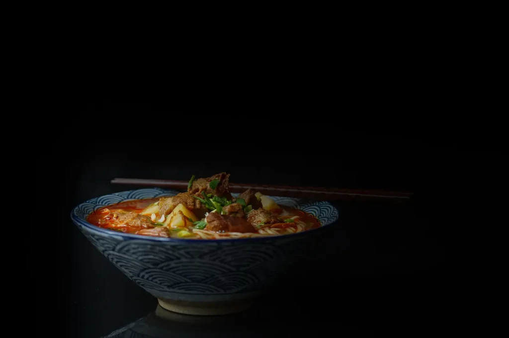 Dark moody food photography of a bowl of noodle soup with rich broth against a black background.