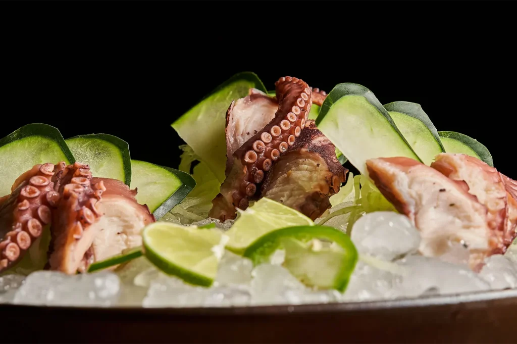 Dark and moody food photography of octopus sashimi served on crushed ice with cucumber slices.