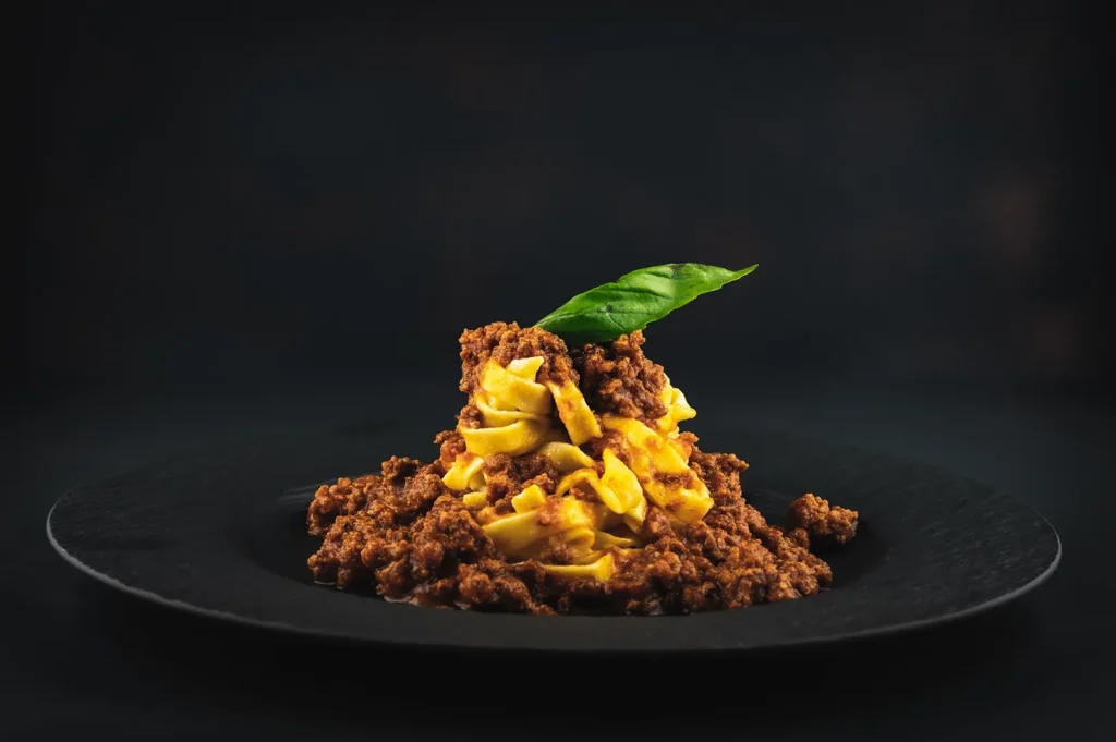 Dark moody food photography of gourmet pasta with meat sauce and fresh basil on a black plate.