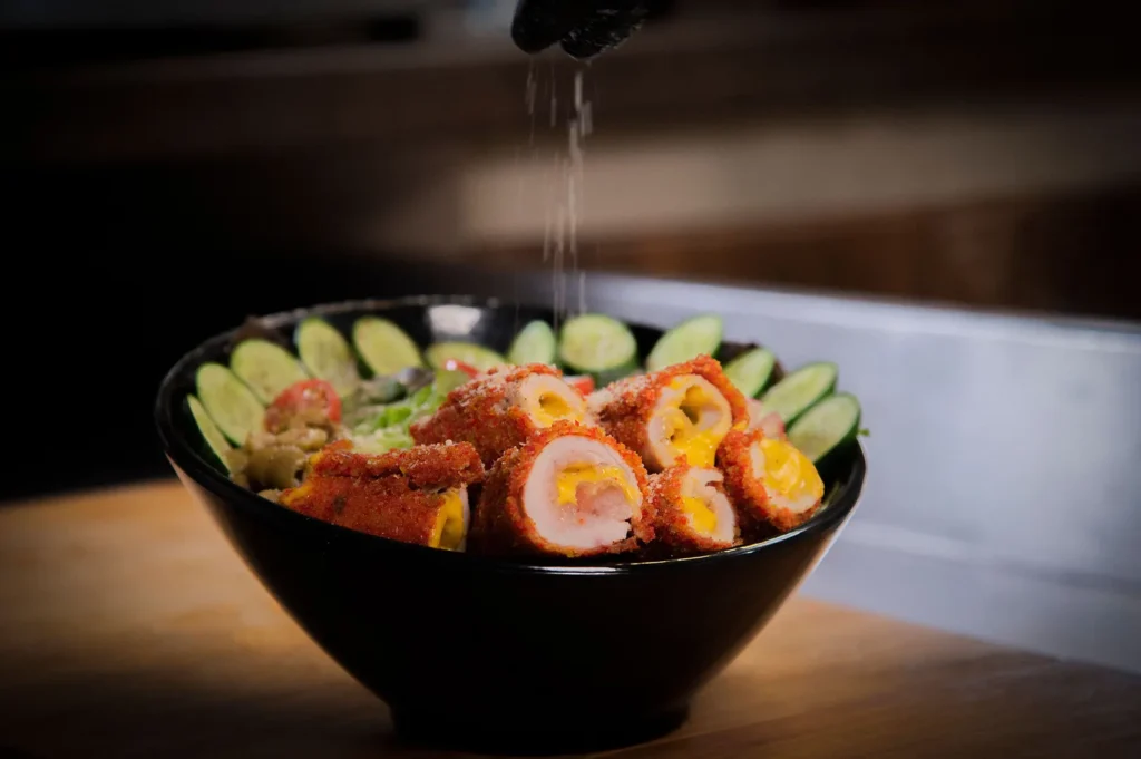 Chef seasoning a gourmet breaded chicken bowl with sliced cucumbers in a dark restaurant setting.