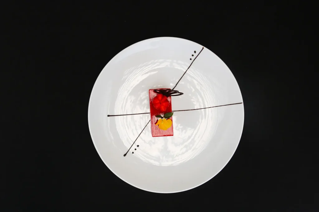 Overhead view of a rectangular red dessert with strawberry, chocolate garnish, and edible flower on a white plate.