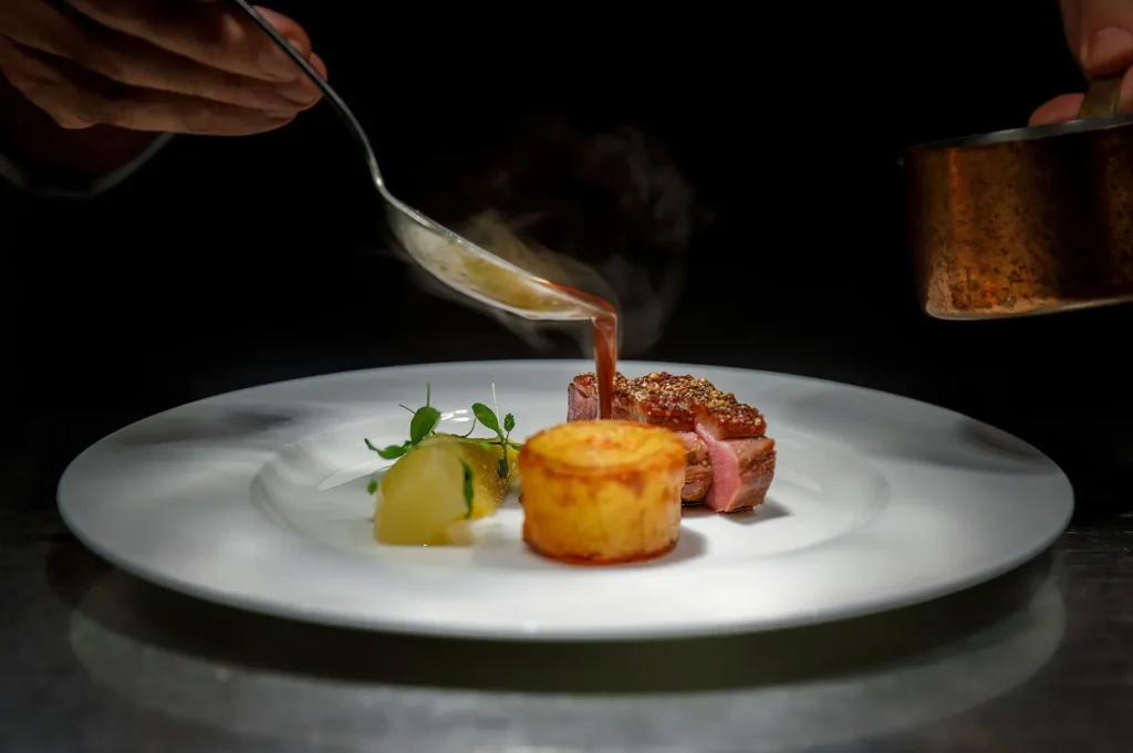 Fine dining hotel room service dish being poured with sauce on a white plate against a dark background.
