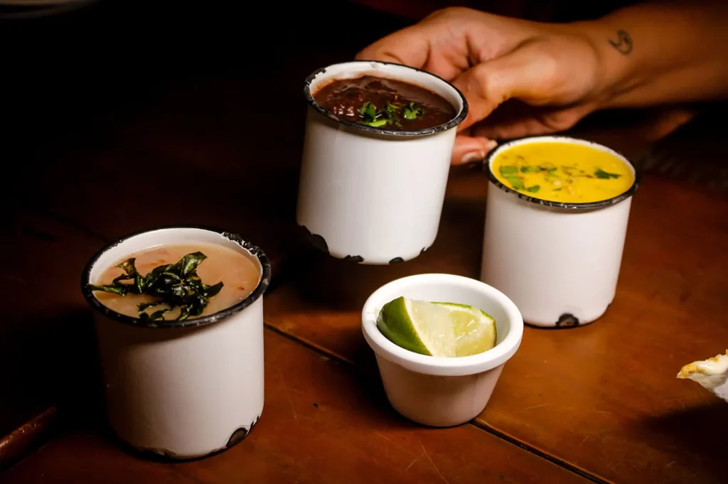 Dark moody food photography of three enamel mugs filled with various dipping sauces and a lime wedge on a dark wooden table.