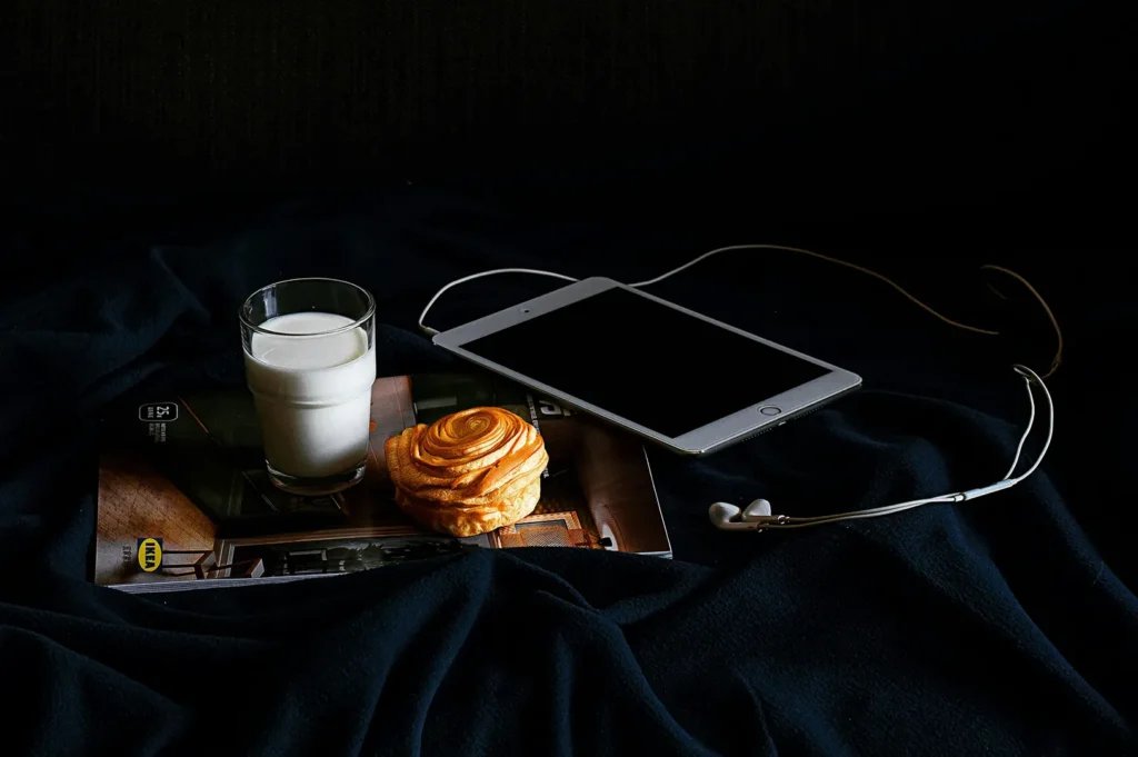 Dark moody food photography styling with a glass of milk, pastry, and digital tablet on a dark background.