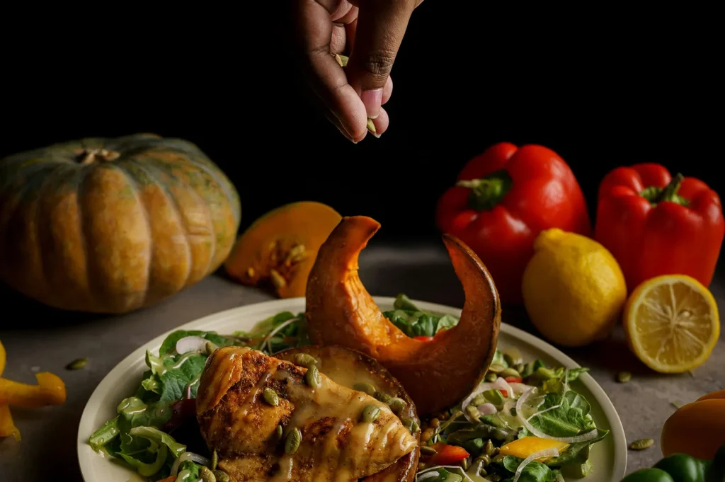 Dark moody food photography of a chef's hand garnishing a roasted pumpkin and chicken salad.