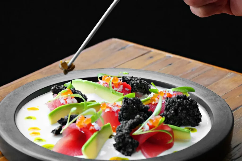 Fine dining gourmet tuna sashimi dish garnished with avocado, caviar, and microgreens on a black plate.