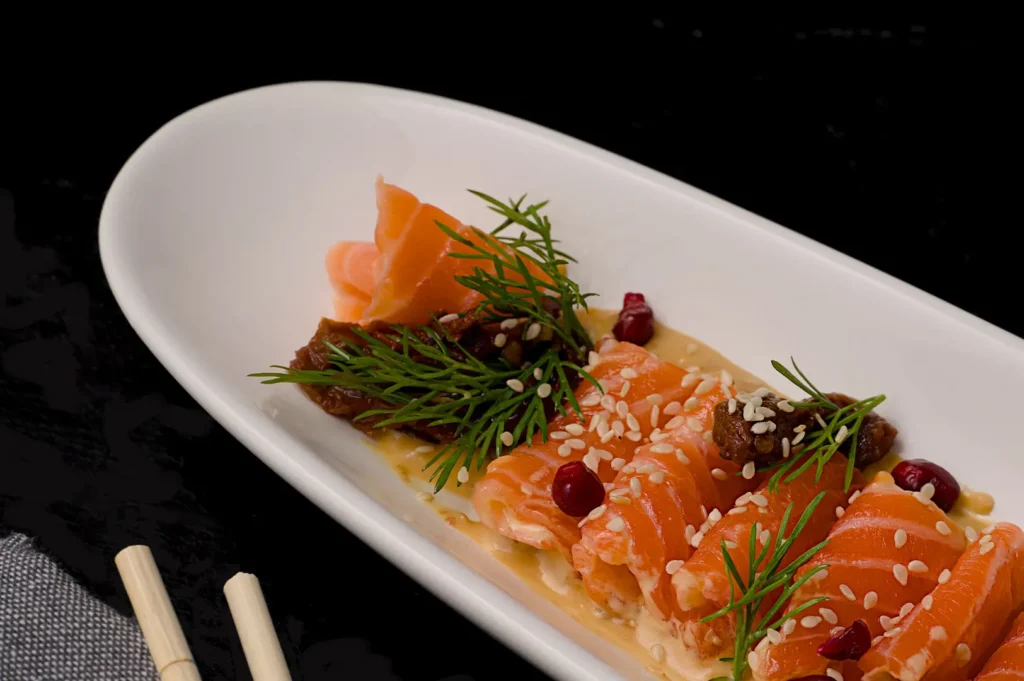 Fresh salmon sashimi plated on a white dish with sesame seeds and herbs.